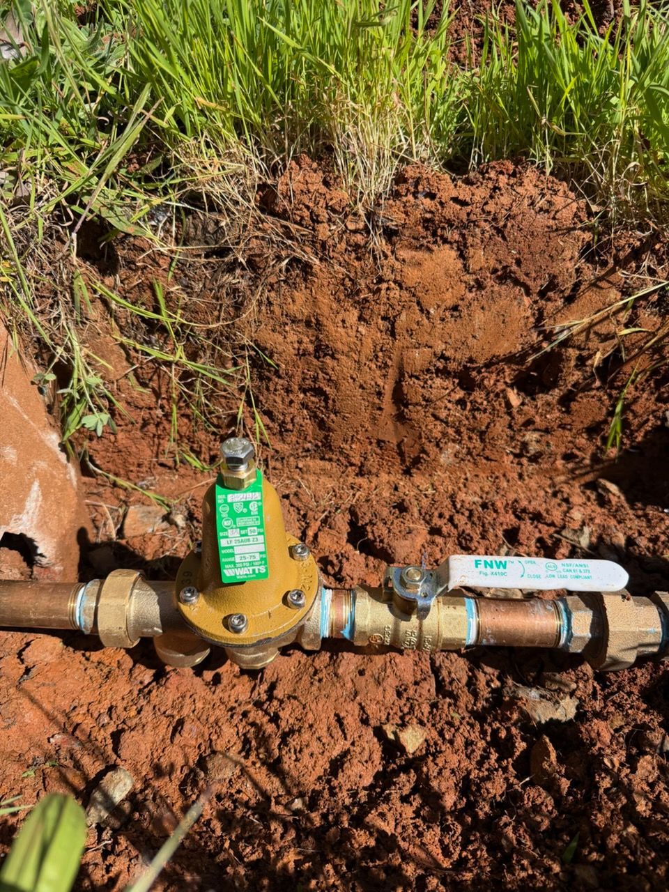 A brass water pressure regulator and a shut-off valve connected to copper pipes, partially buried in red soil.