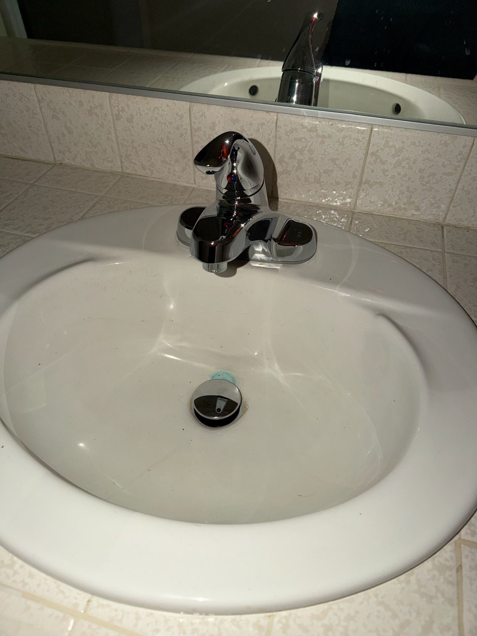 A clean, white bathroom sink with a polished chrome faucet, set into a tiled countertop with a mirror in the background.