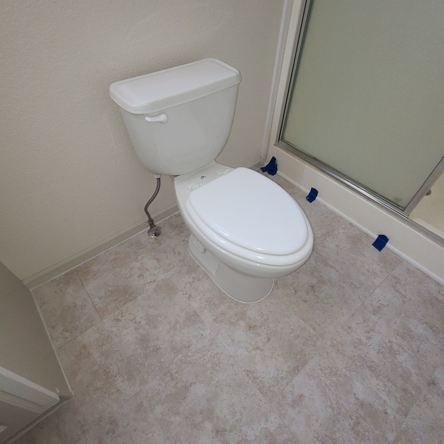 Toilet in a bathroom with light-colored flooring. It's positioned next to a shower with a glass door.