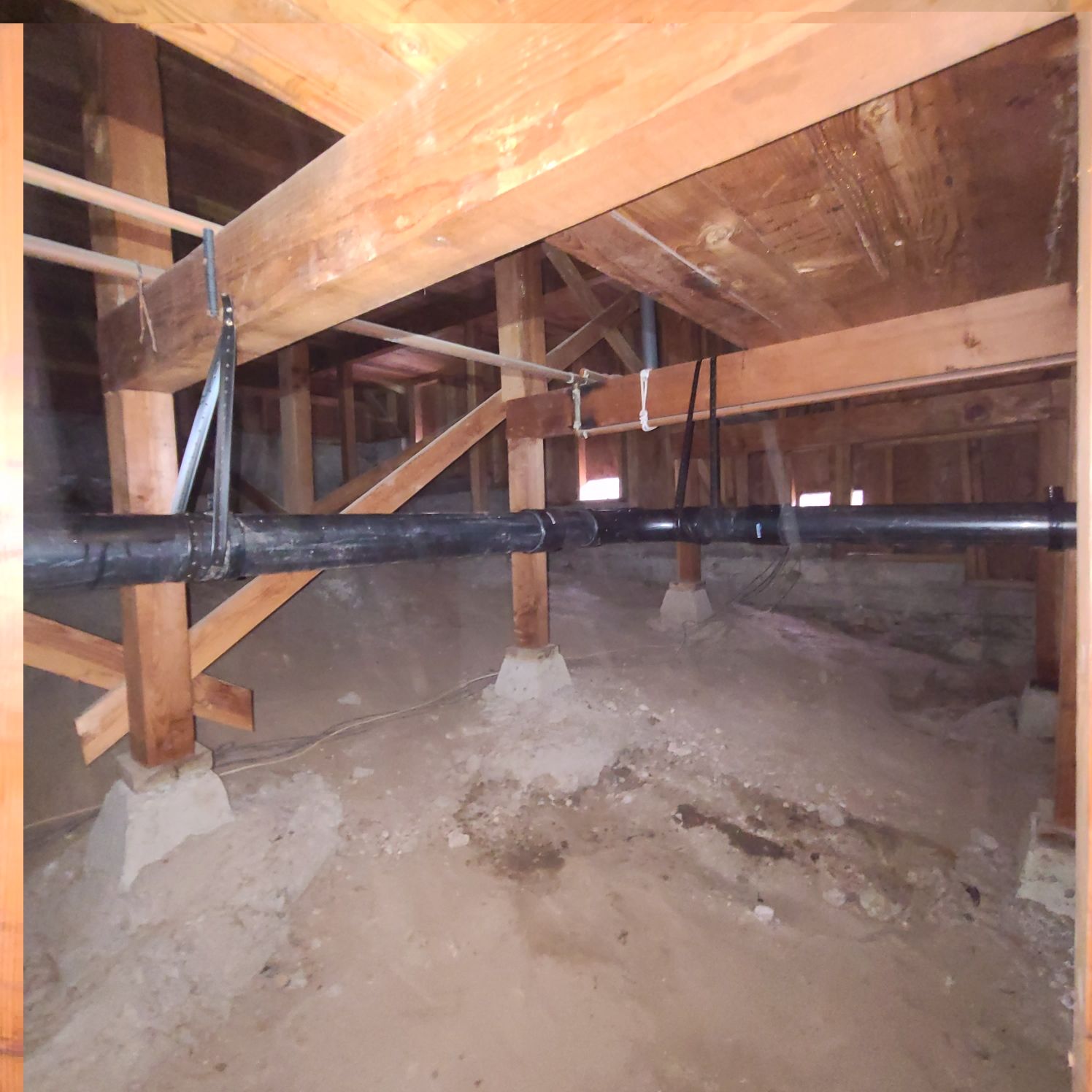View of a crawl space with wooden beams, support posts, and a black pipe suspended from above.