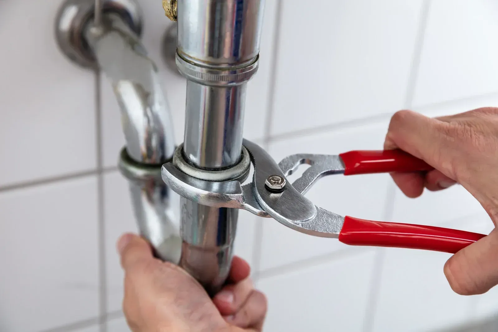 Hands using pliers to tighten a silver pipe under a sink.