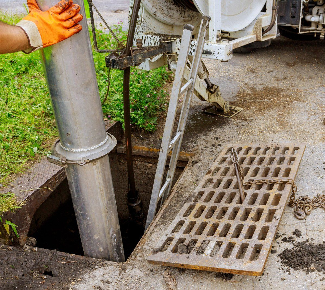 A person in orange gloves lowering a large metal pipe into an open sewer manhole on a concrete street.