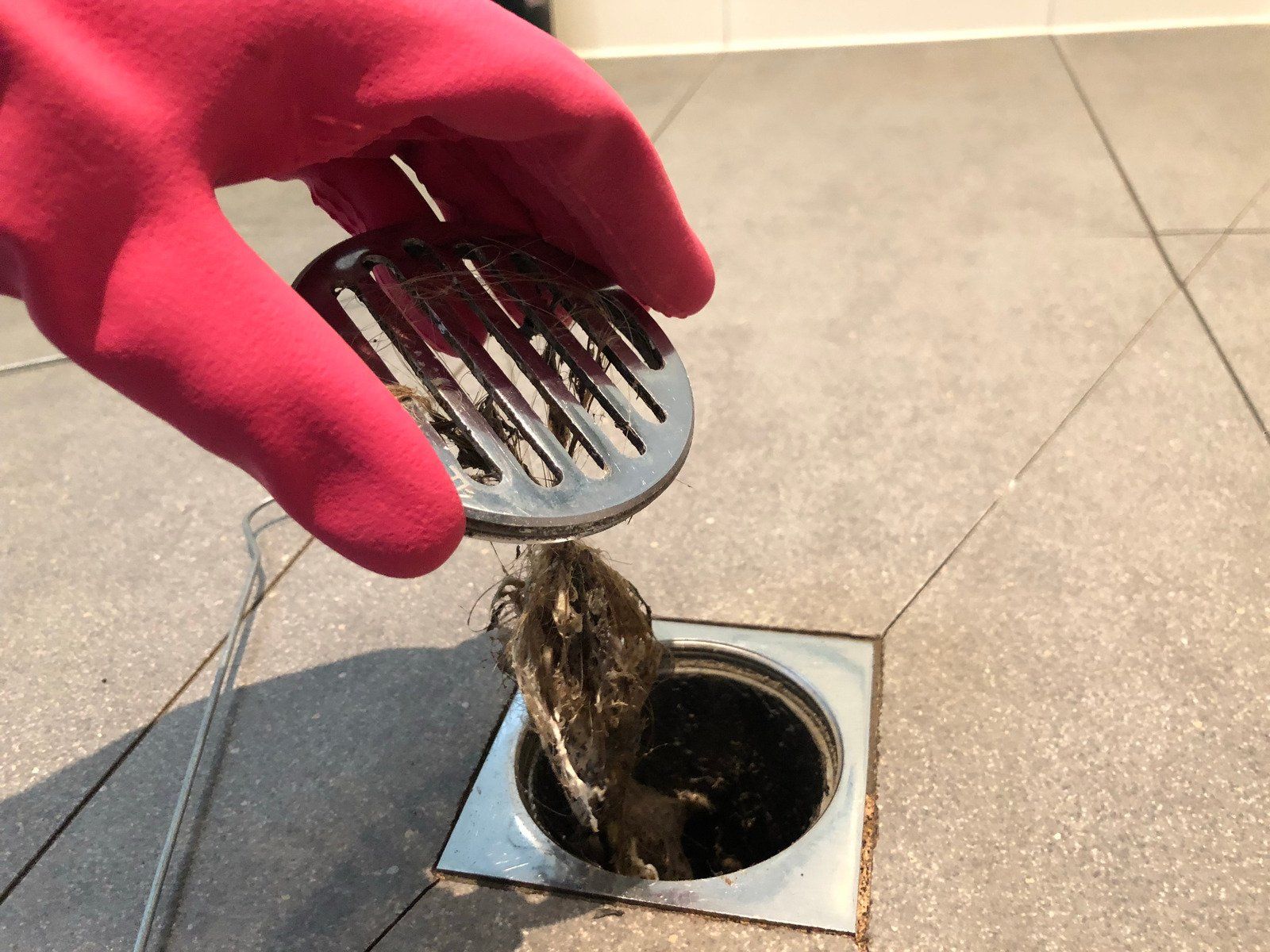 A gloved hand lifts a drain cover, revealing a clogged shower drain filled with hair and debris.