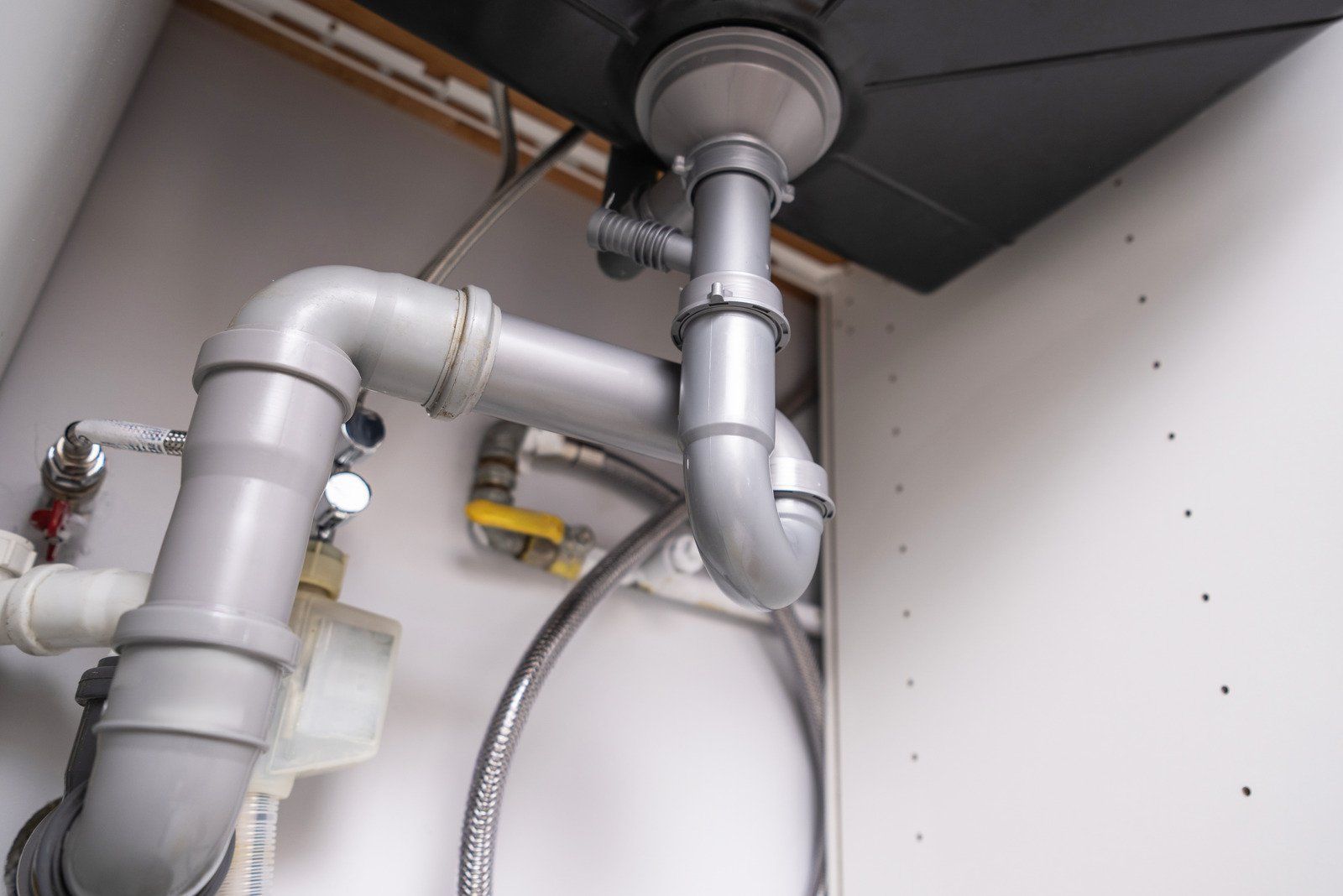 Plumbing pipes beneath a kitchen sink, with a curved drainpipe and various connecting pipes in a white cabinet.
