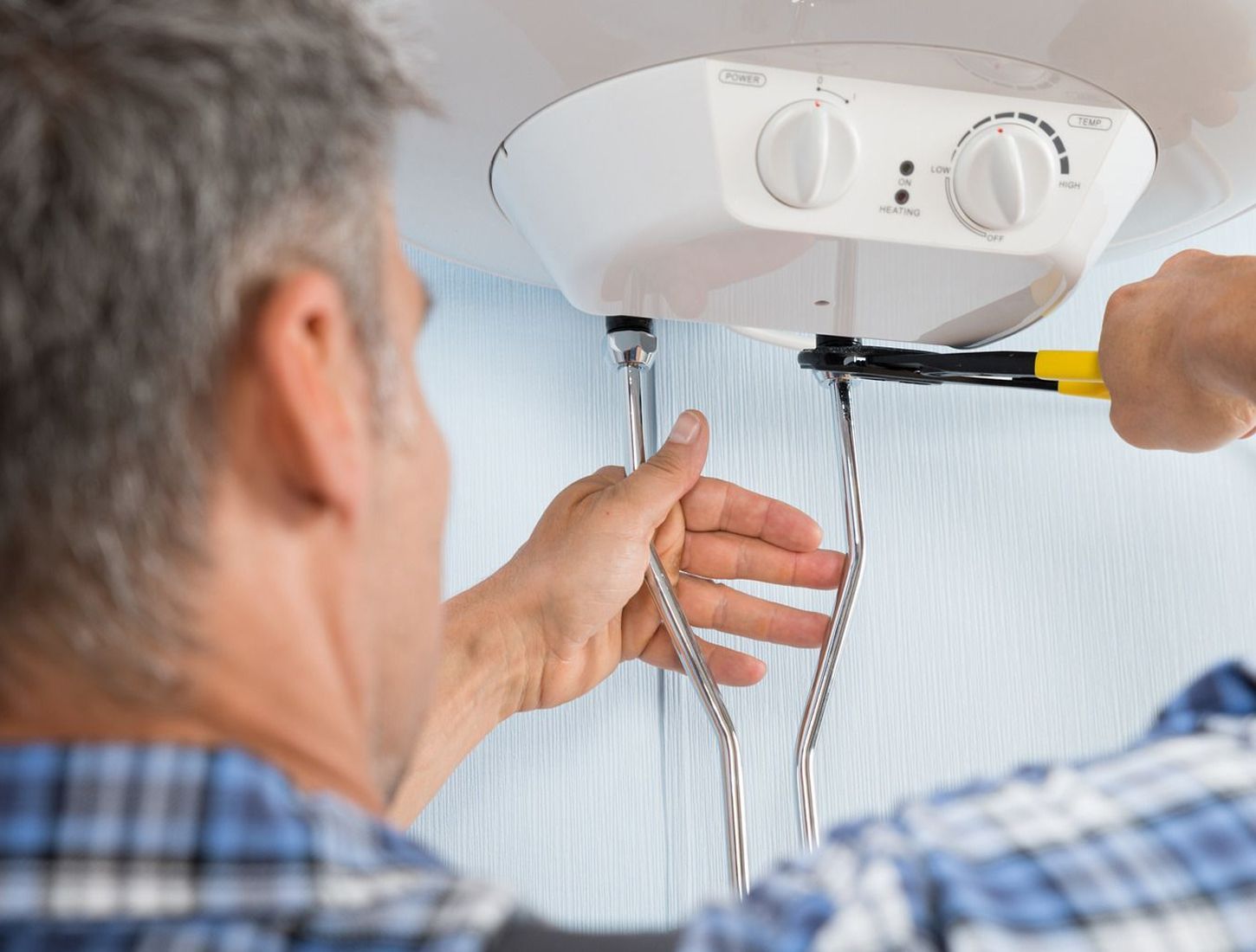 Man in plaid shirt using pliers to work on a white water heater mounted on a light blue wall.