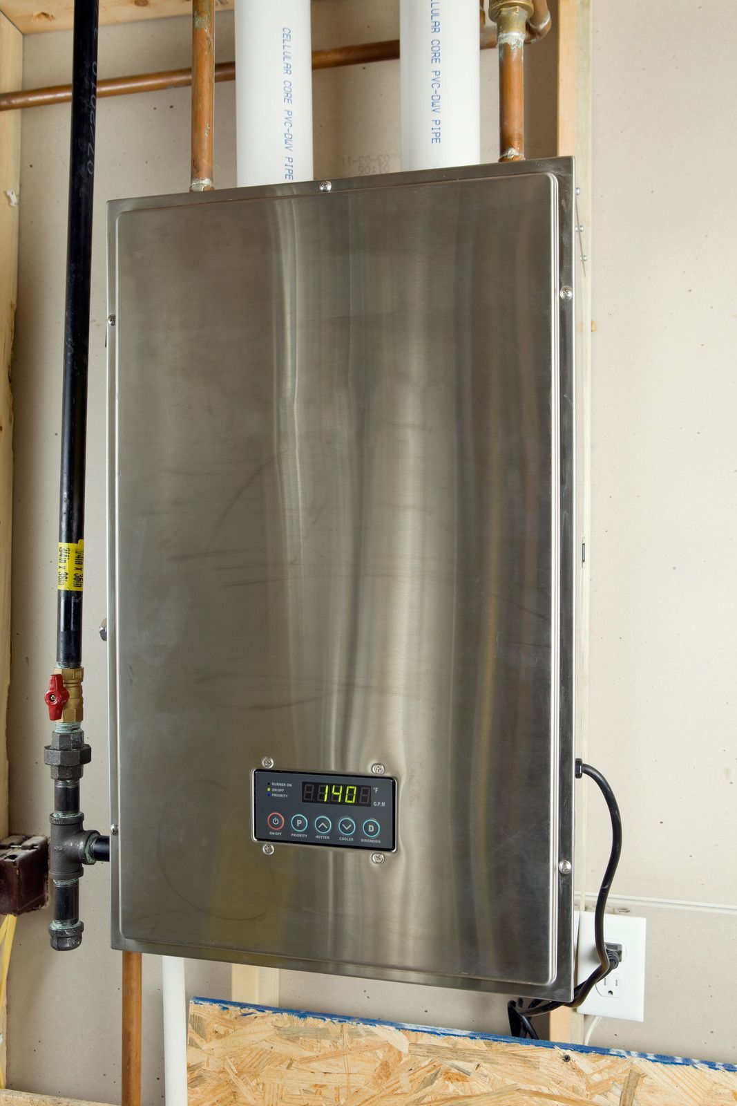 A stainless steel tankless water heater mounted on a wall with pipes and a black gas line.