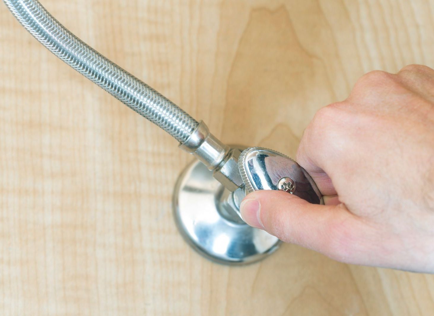 Hand turning a chrome water shut-off valve attached to a flexible supply line against a light wooden surface.
