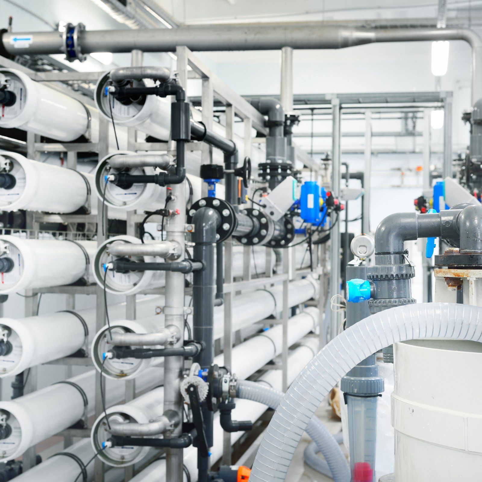 Industrial water treatment plant with white cylindrical filter housings, pipes, valves, and a gray corrugated hose.