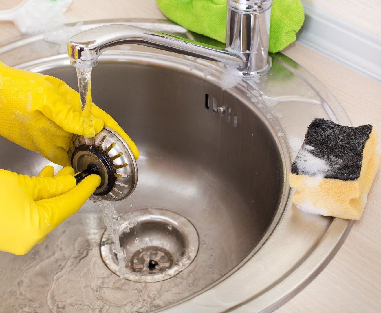 Hands in yellow gloves cleaning a kitchen sink drain with a strainer under running water. A sponge sits nearby.