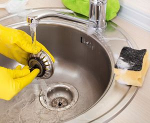 Hands in yellow gloves cleaning a kitchen sink drain with a strainer under running water. A sponge sits nearby.