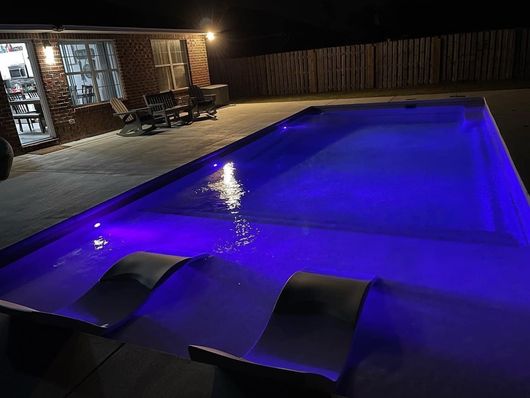 A large swimming pool is lit up at night with purple lights.