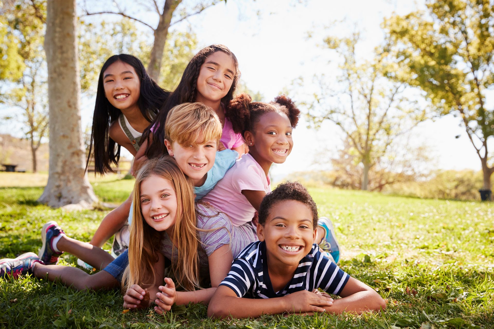 A group of children are laying on top of each other in the grass in a park.