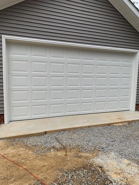 White garage door with grid pattern, set in white trim, against gray siding, on a concrete base with gravel.