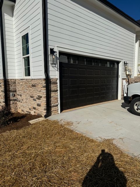 Black garage door on a white and brick house exterior. Concrete driveway and dry grass in front.