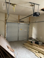 Interior of a garage with a partially open silver garage door, building materials on floor, exposed ceiling.