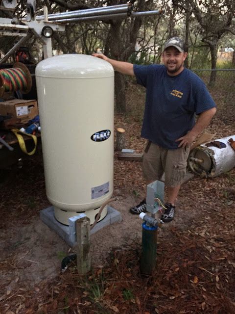 A person stands next to a large cream-colored well pressure tank installed outdoors on a concrete base.