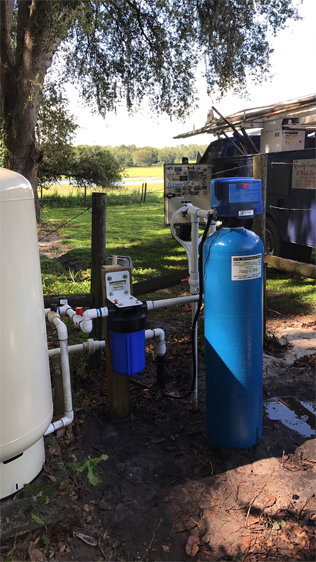 A white well pressure tank connected by pipes to a blue water filter and a blue water treatment tank in an outdoor yard.