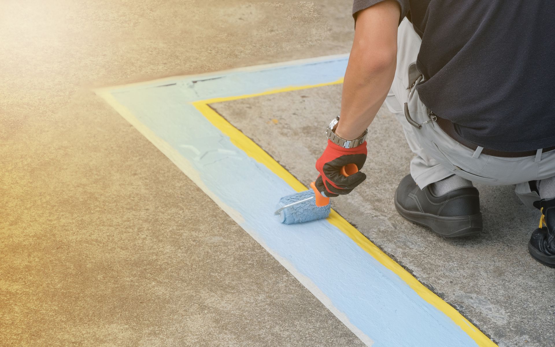 A man is kneeling down and painting a parking lot with a paint roller.