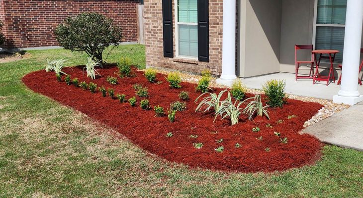 A garden with red mulch and plants in front of a house.