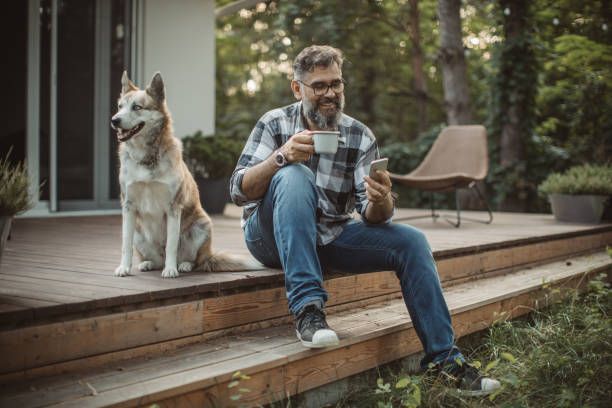 Man sitting on deck with a dog, holding a coffee mug and phone. Outdoors with trees in the background.