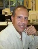 Man in a light-colored shirt smiles, resting his hand near his face. He is seated in an office with papers and a computer.