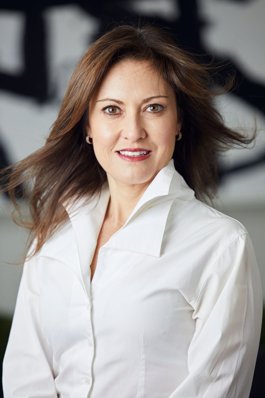 Woman in a white shirt smiles, hair blowing, against a blurred art backdrop.