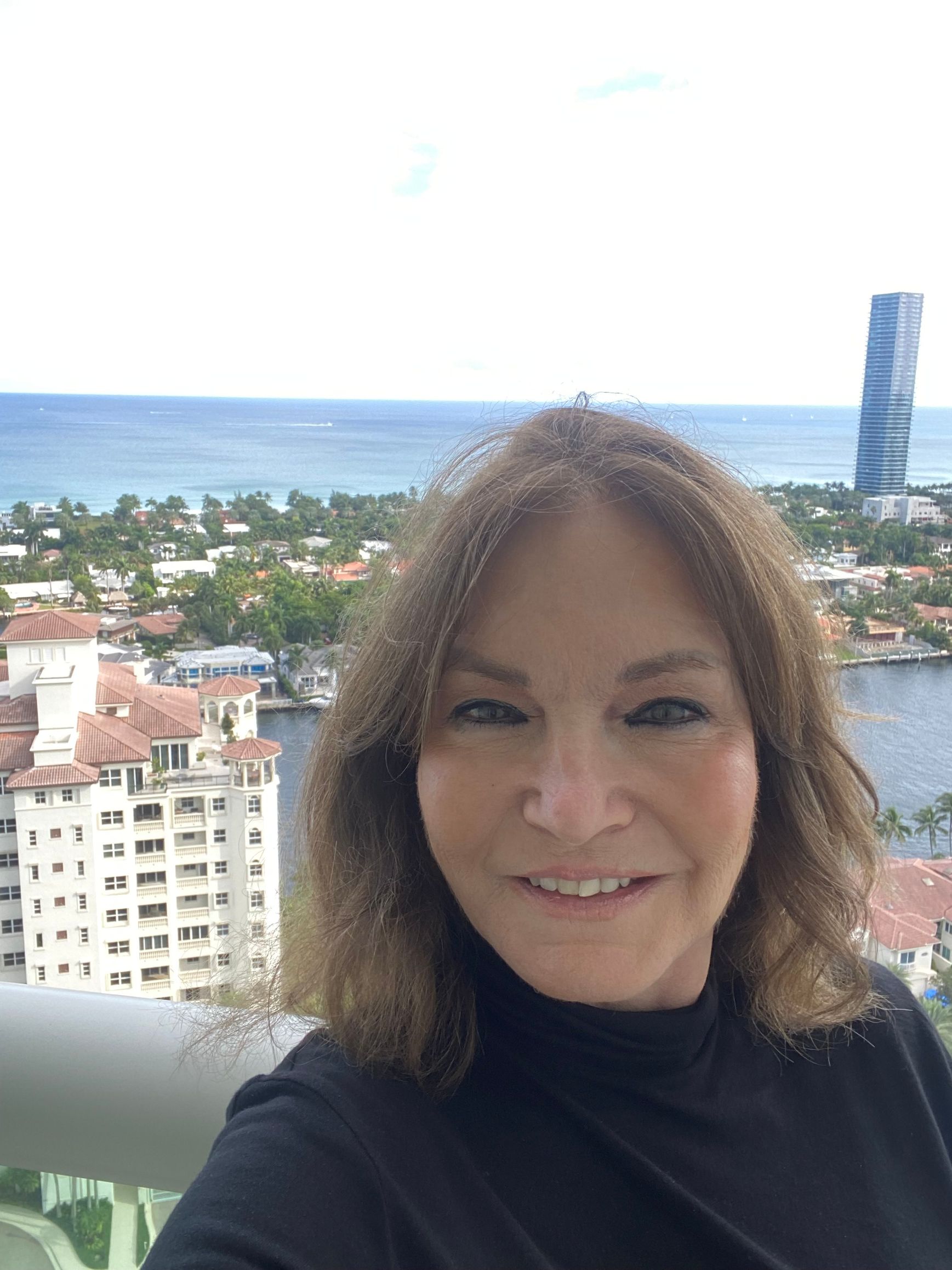 Woman smiles in front of a waterfront view. She wears black, with brown hair. Sunny outdoors.