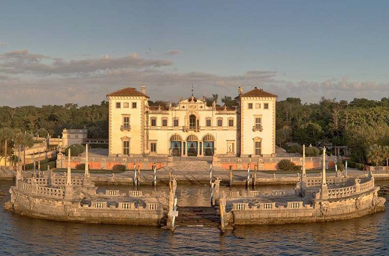 An ornate, light-colored Italian Renaissance style villa on a waterfront platform, surrounded by water.