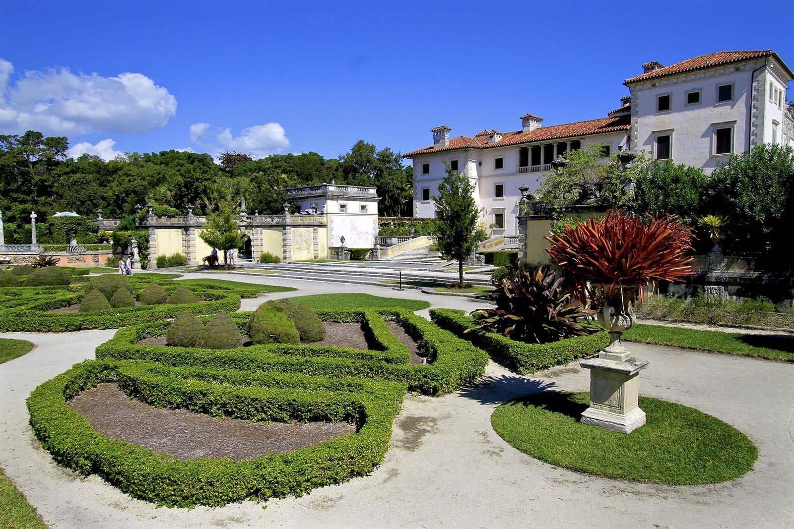 Formal garden with hedge rows, gravel paths, and a large white villa under blue sky.