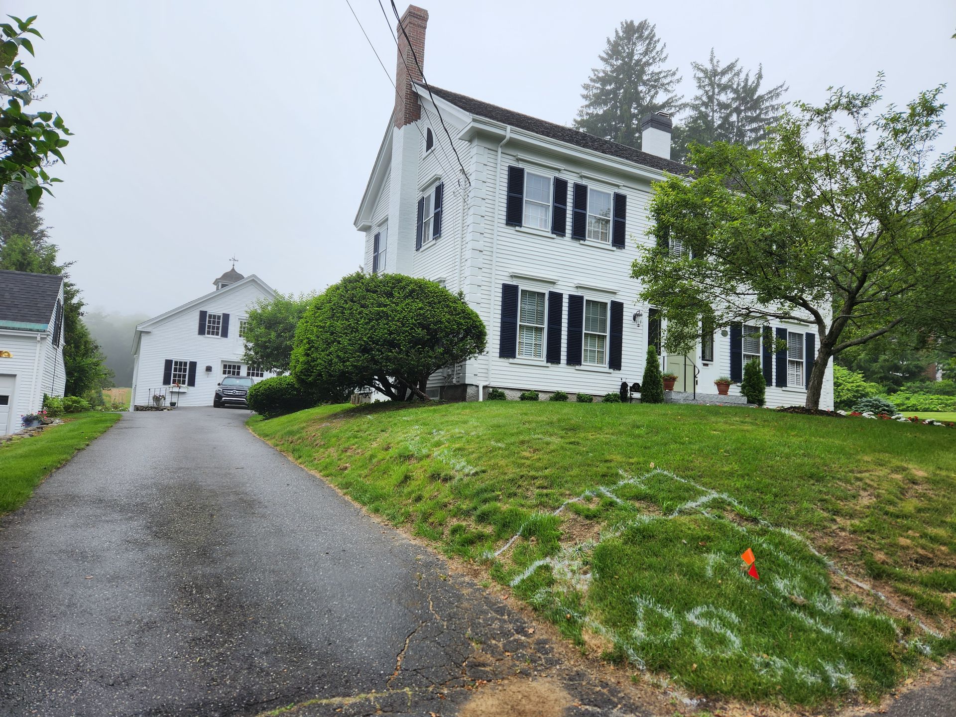 White two-story house with black shutters, driveway, and grassy yard on a cloudy day.