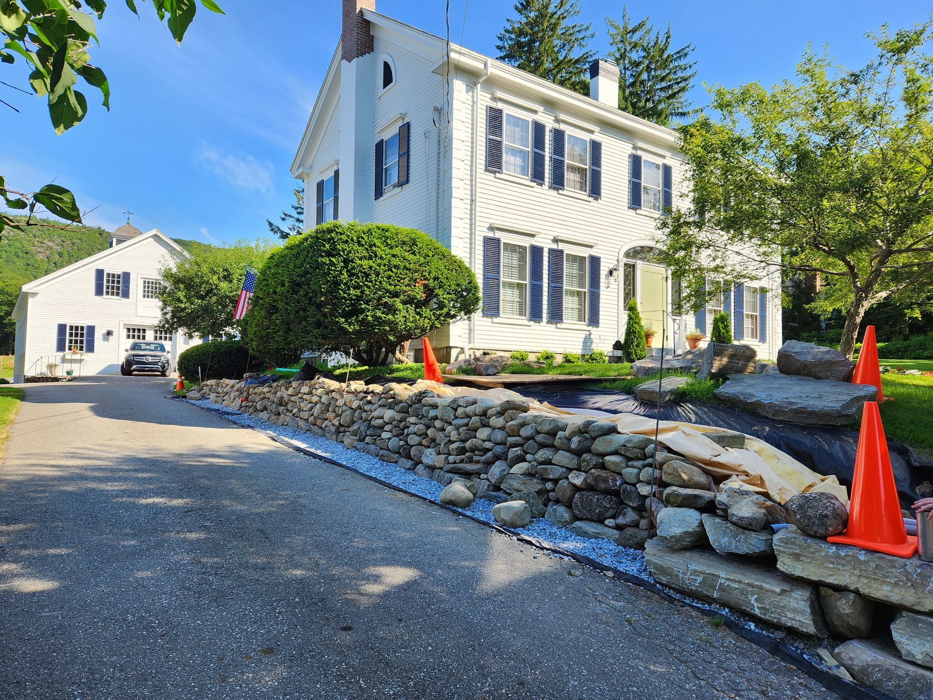White house with blue shutters, stone wall, and driveway on a sunny day.
