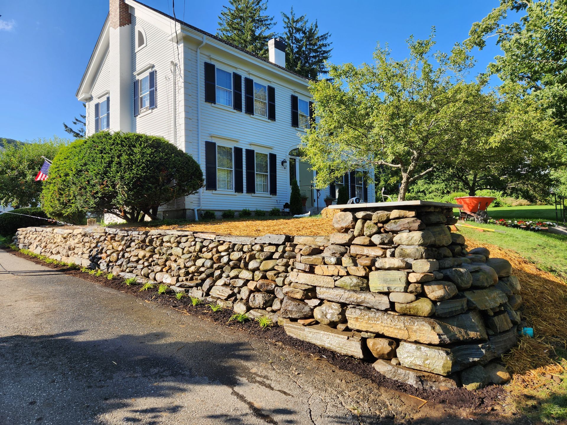 White two-story house with black shutters, stone wall, and green trees on a sunny day.