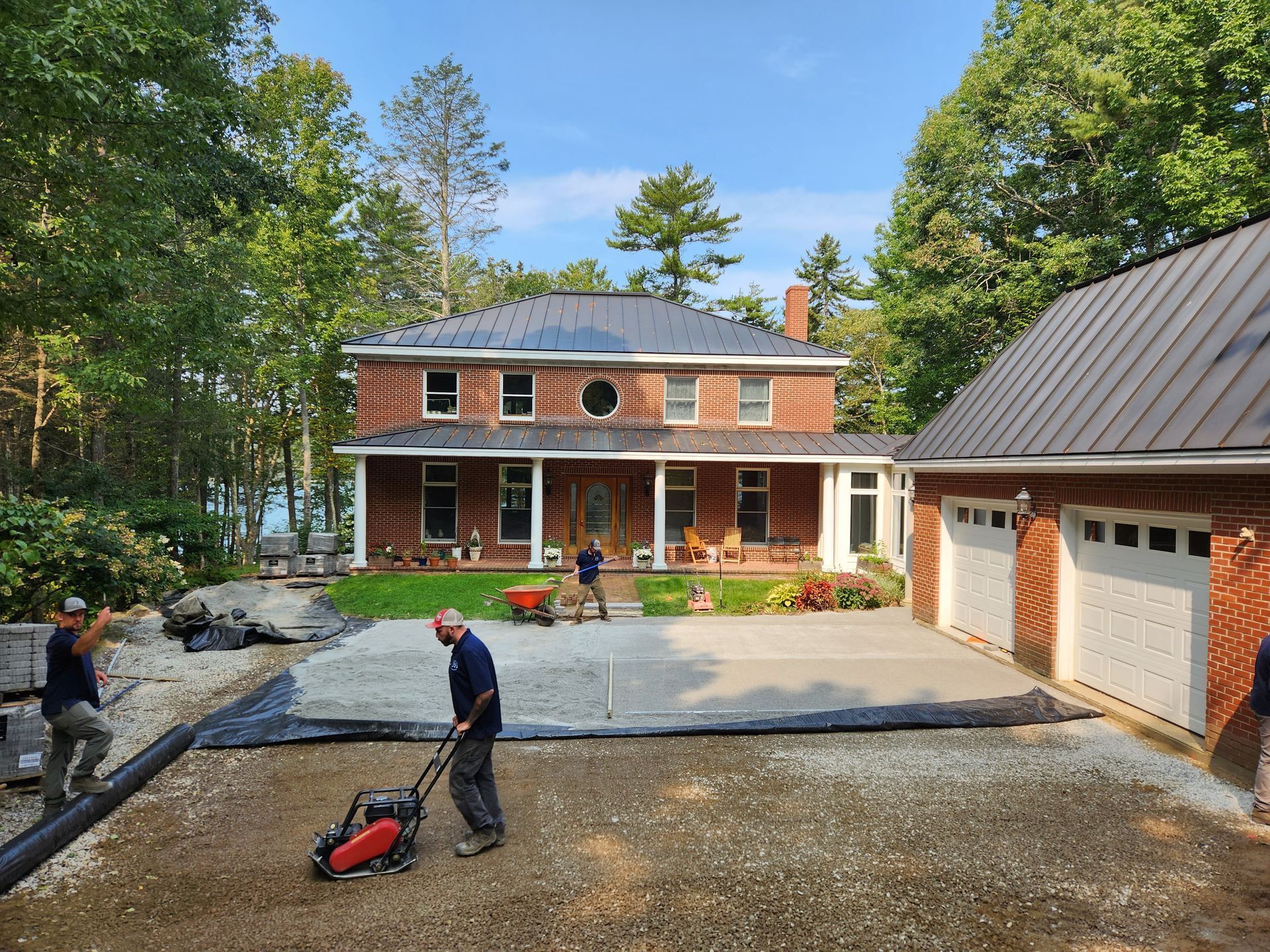 Construction workers leveling gravel driveway near a brick house, a detached garage, and trees under a blue sky.