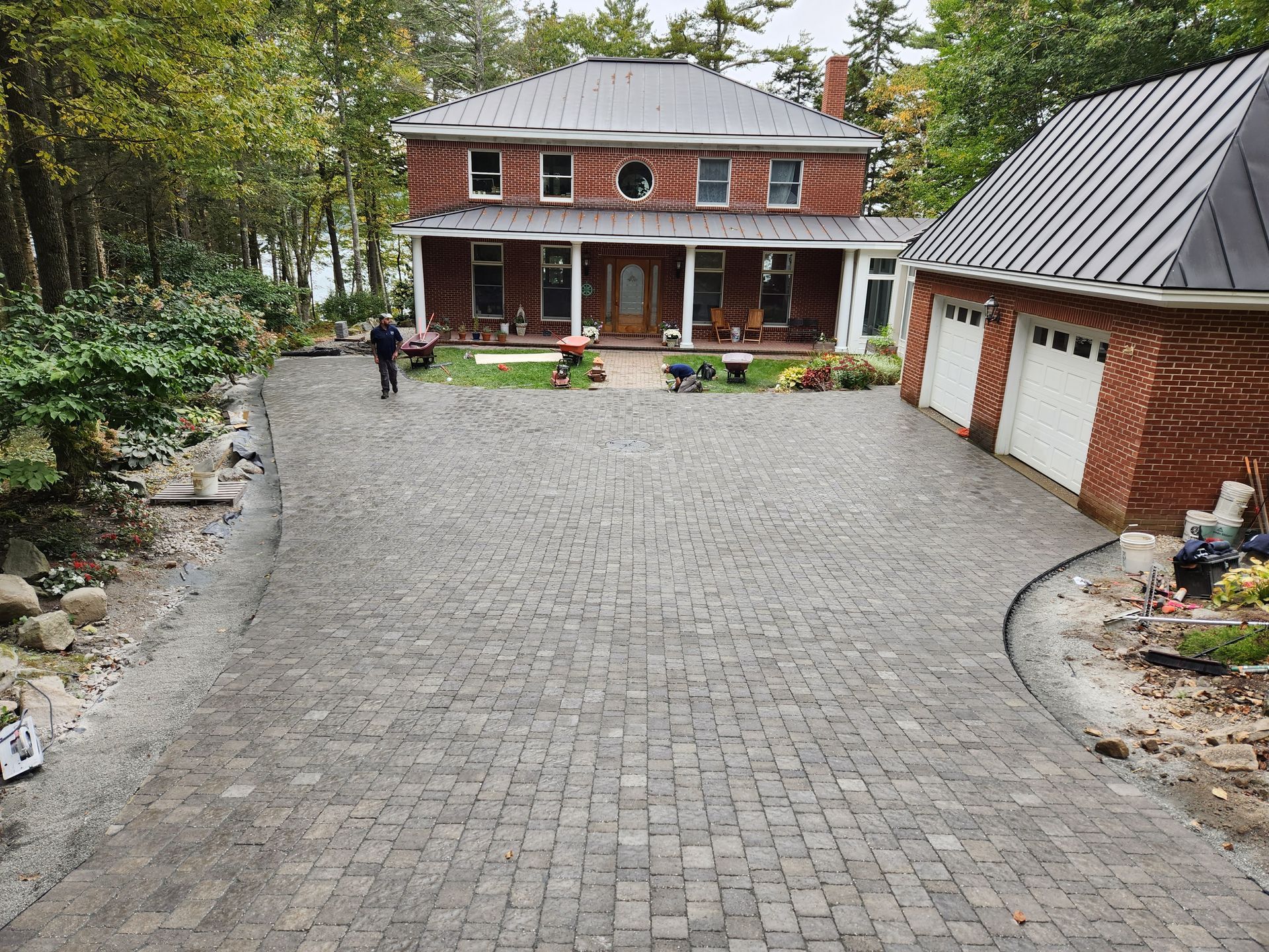 Brick home with brick driveway. Person walks towards the house. Garage on the right. Trees in the background.