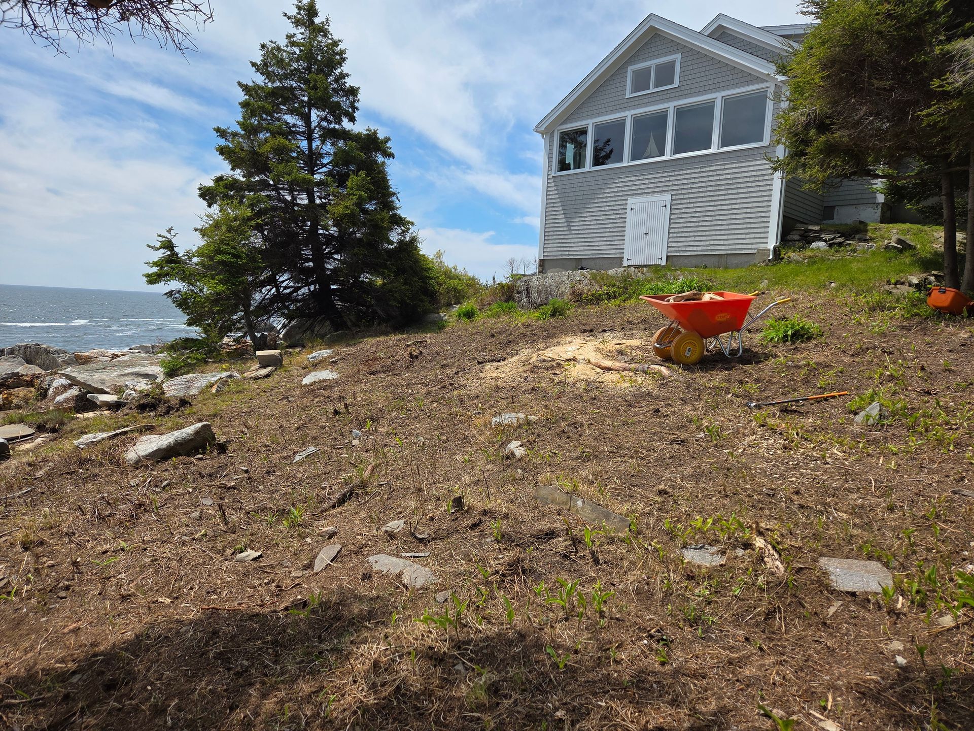 Coastal house with ocean view; person using spreader on grassy hillside, blue sky.