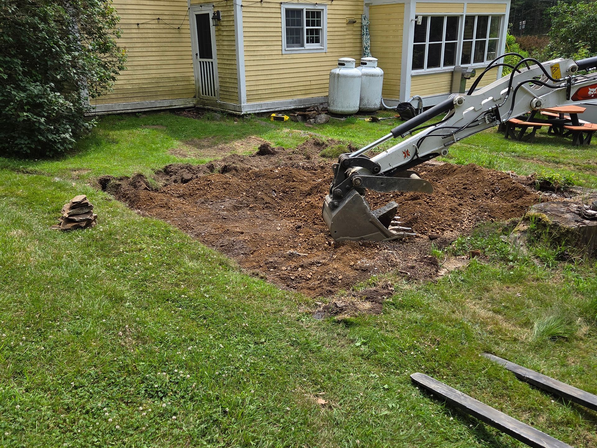 A small excavator digs into a patch of dirt in a grassy yard, near a yellow house.