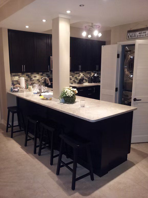 Kitchen with dark cabinets, a white countertop island with bar stools, and a laundry room door.