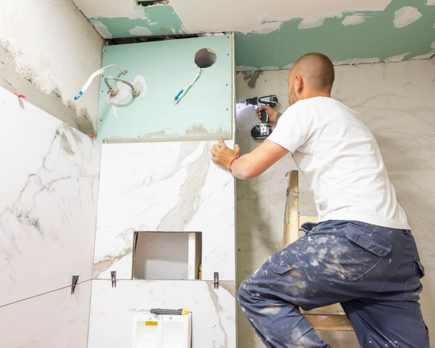 Man installing tiles in a bathroom, using a drill. He's on a ladder, working on a wall with plumbing visible.
