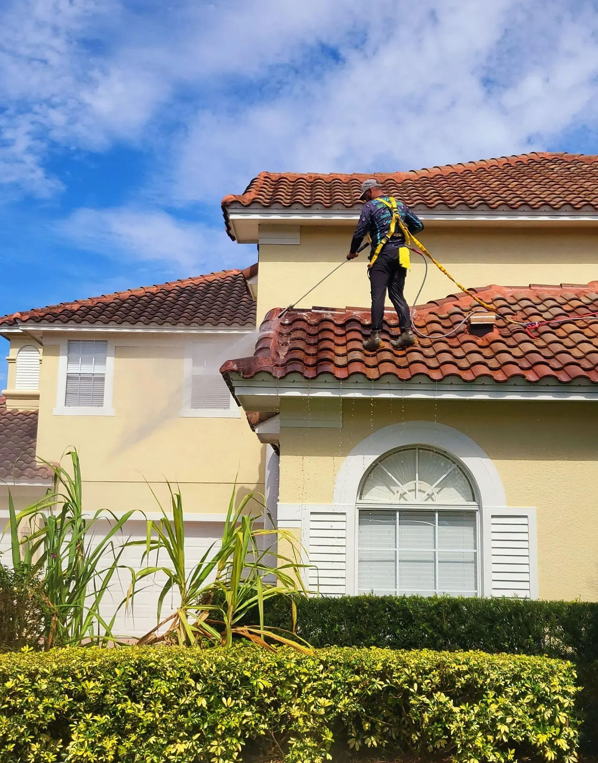 A worker in safety gear pressure washing a red tile roof on a yellow house under a blue sky.