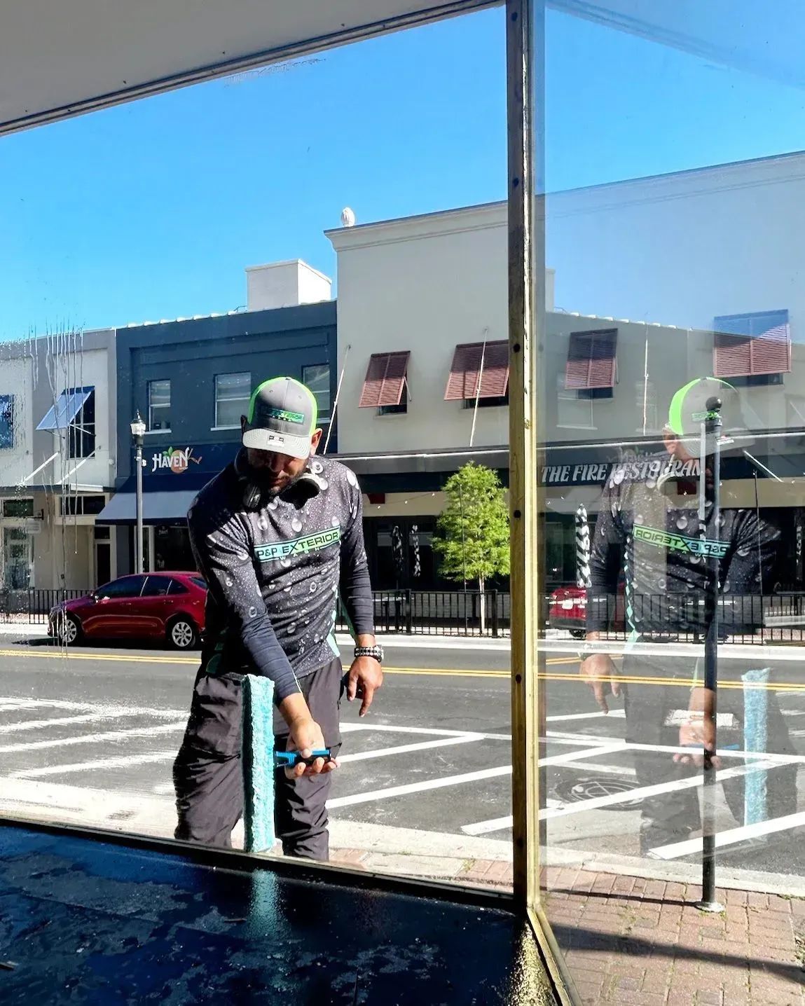 A person washing a large store window with a cleaning tool, downtown setting with storefronts, and a red car.
