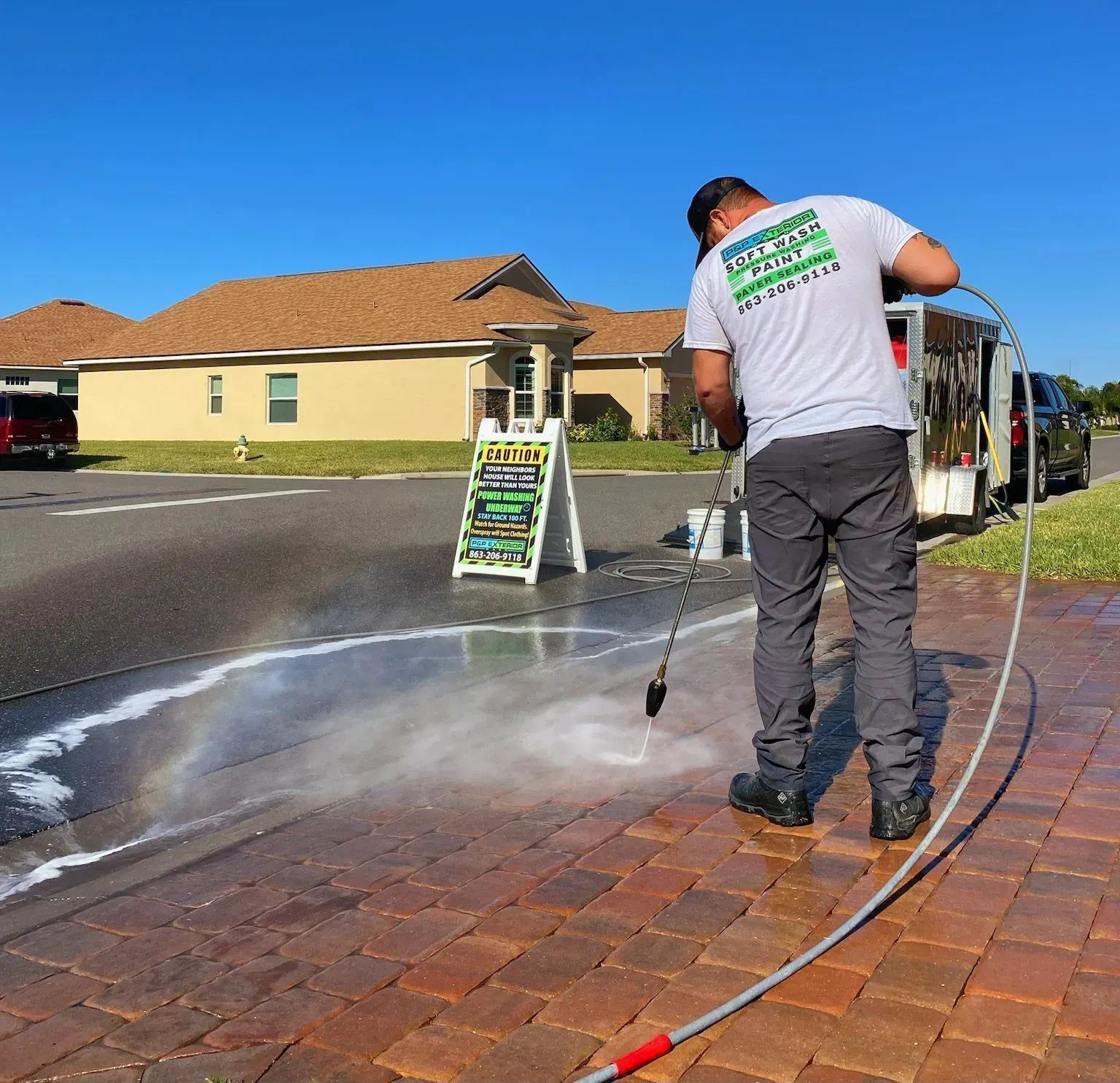 A worker in a gray t-shirt pressure washes a brick paver driveway near a sign on a sunny day.