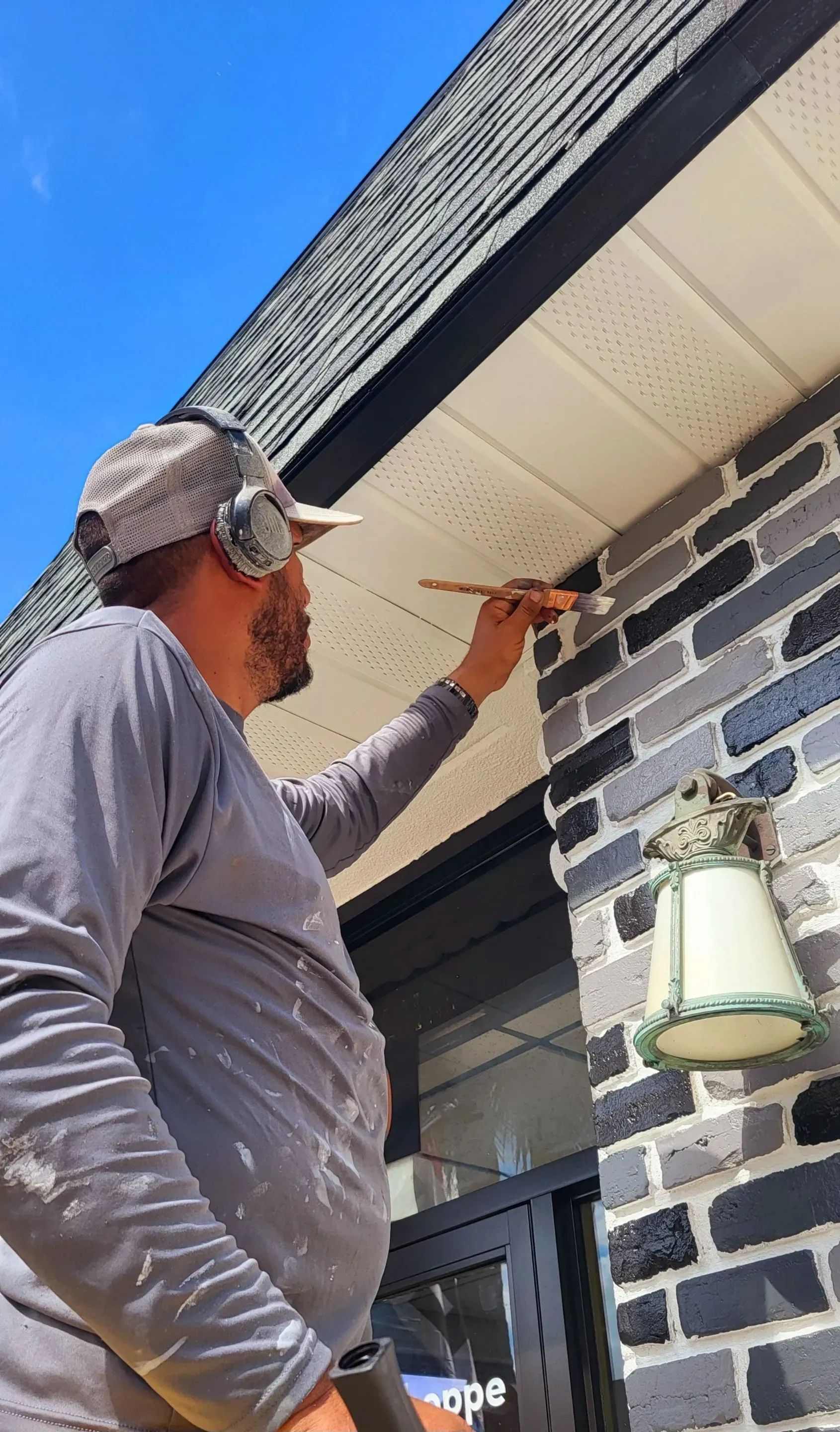 Person painting trim on a building's exterior, wearing a cap and ear protection.