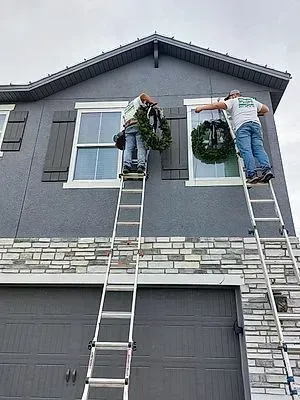 Two men are standing on ladders decorating a house with a wreath.