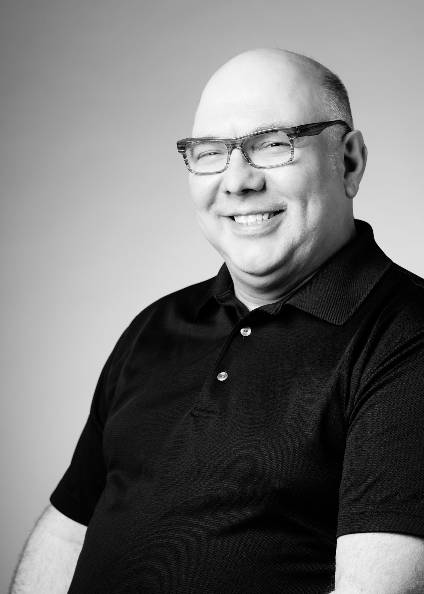 Homme souriant et chauve portant des lunettes et un polo noir.