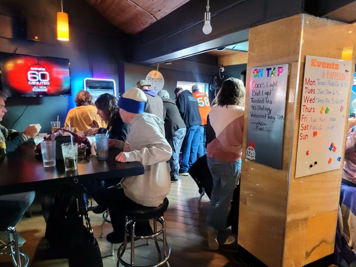 People chatting in a dimly lit bar, with colorful neon signs and handwritten menu boards.