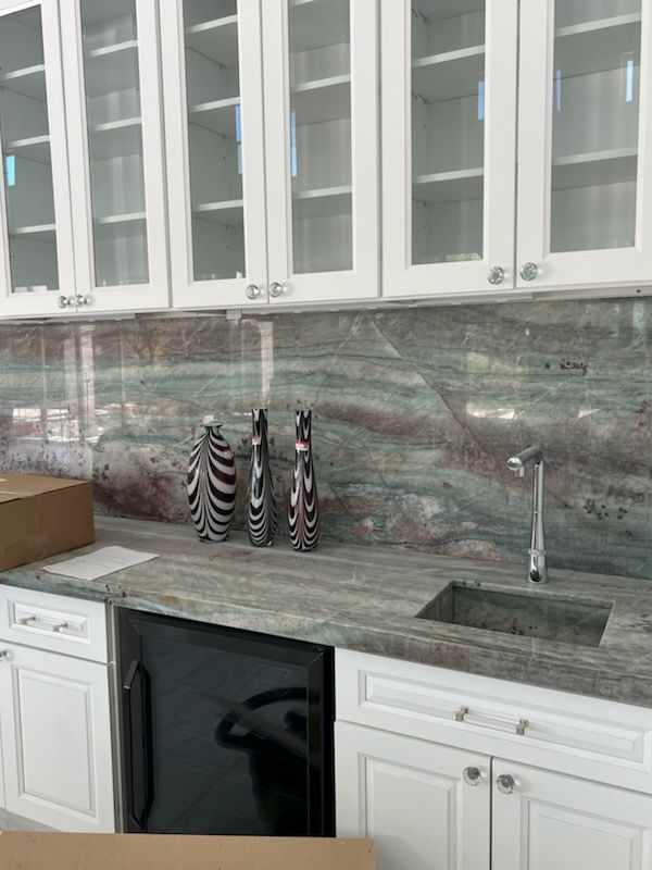 White cabinets with glass doors over a green and brown granite backsplash, sink, and mini-fridge.