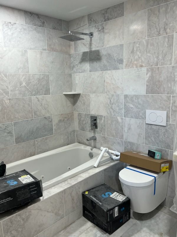Bathroom with gray tiled walls, bathtub, showerhead, and a white toilet. Boxes on the floor.