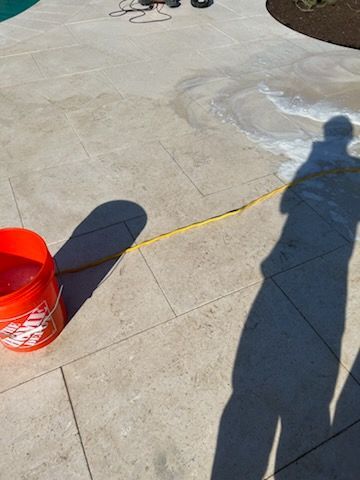 Person cleaning a light-colored stone patio with a power washer. A bucket of soapy water and a yellow cord are visible.