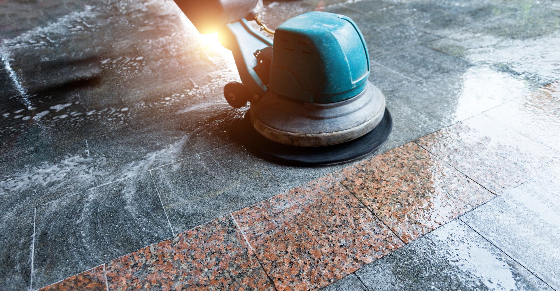 Floor polishing machine in use on a granite floor, reflecting light and water.