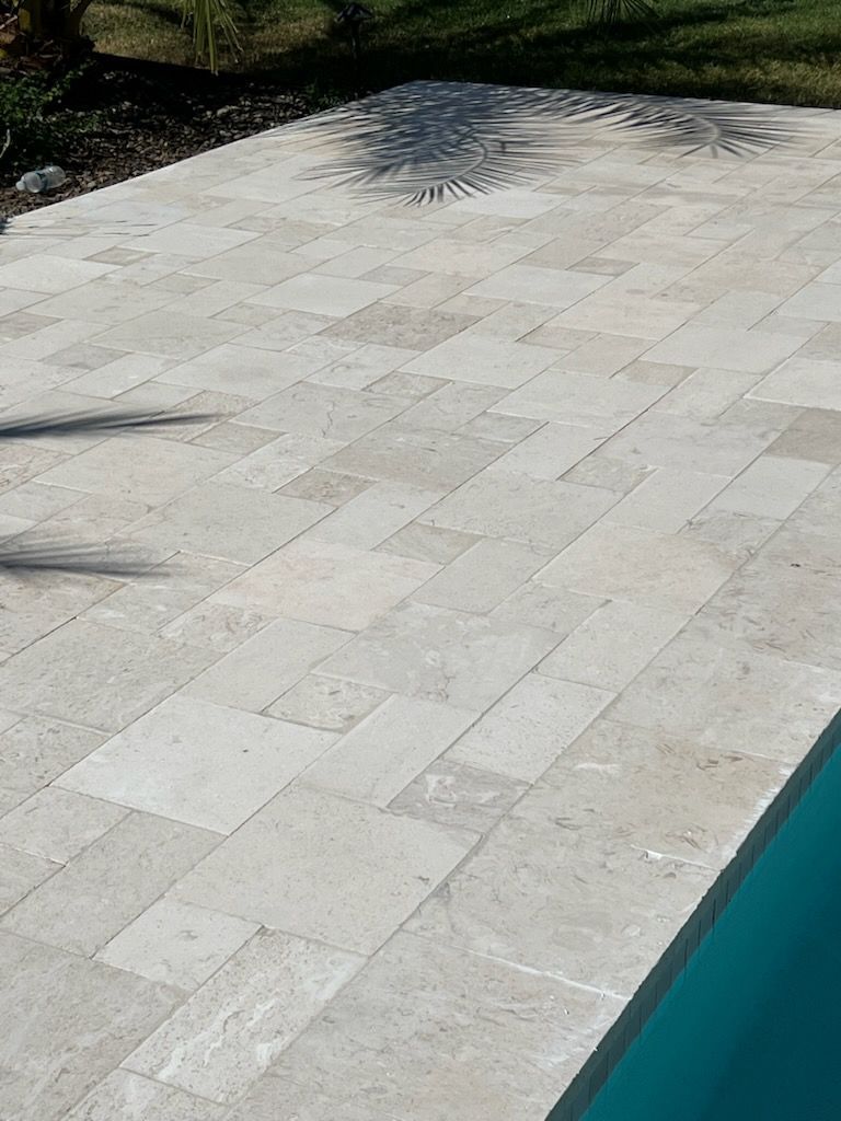 Beige stone patio next to a turquoise pool. Sunlight and shadows highlight the textured stone.